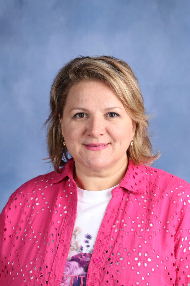 Maia Patic, smiling woman with blonde hair wearing a pink shirt against a blue background.