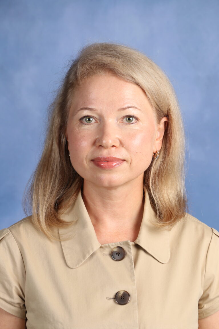 Liudmila Rakhmanova portrait; blonde woman in a tan jacket against a blue background.