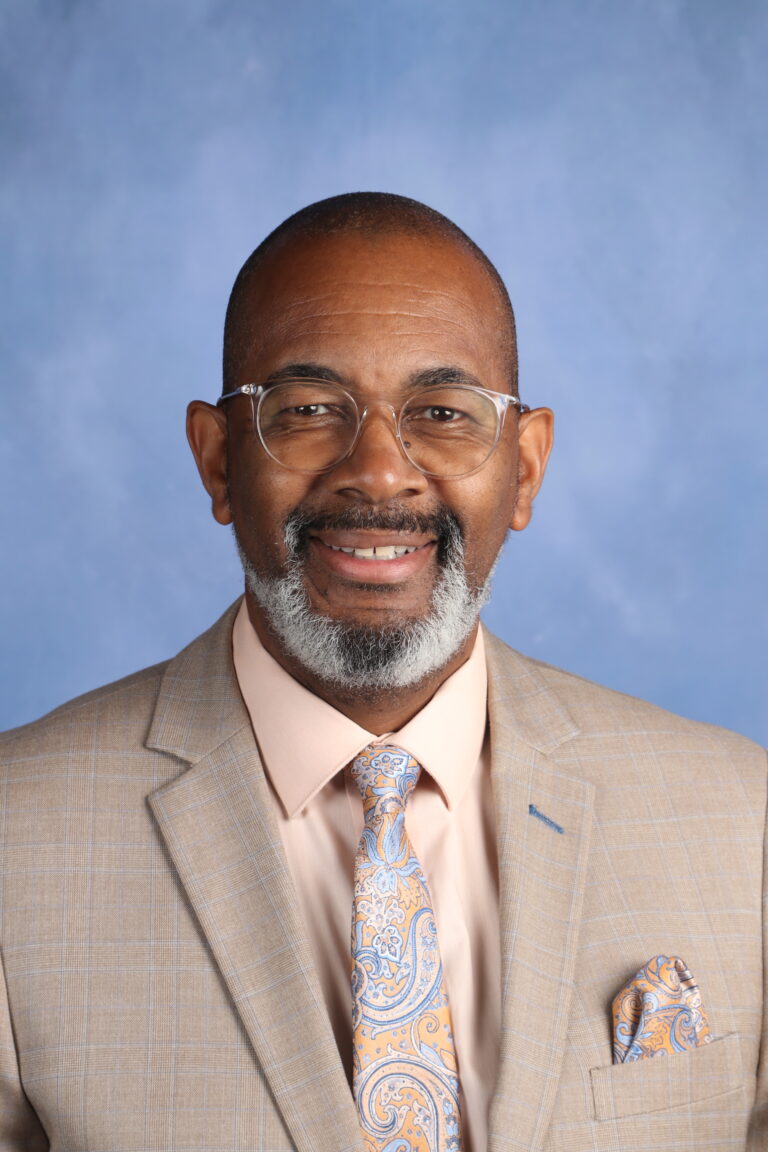 Anthony Brodie, smiling man in suit and glasses against a blue background.