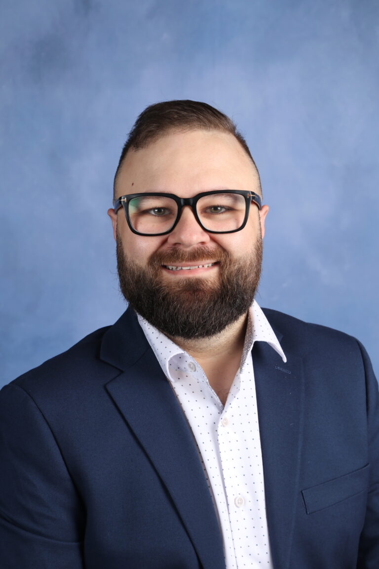 Alex Dragu portrait. Man with glasses and beard in a blue suit smiles for the camera.