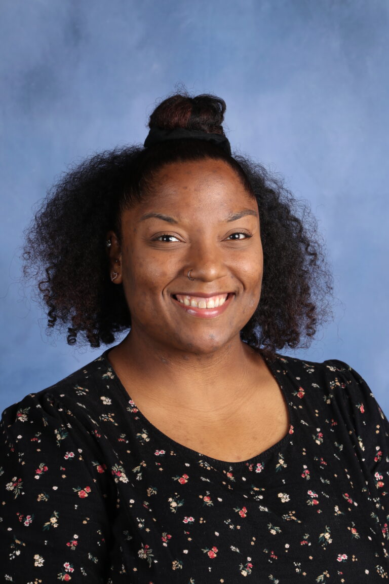 April Webb portrait. Smiling woman with curly hair and a floral top against a blue background.