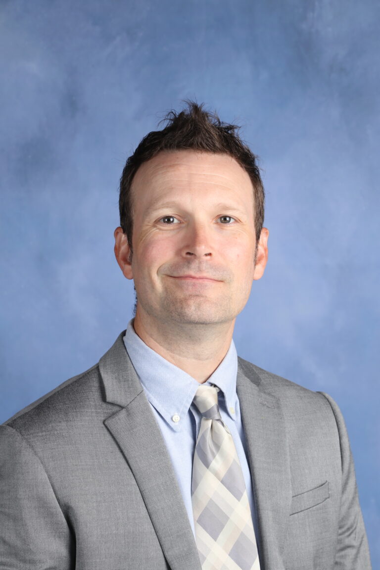 Clayton Shaw in a gray suit and light blue shirt, smiling.