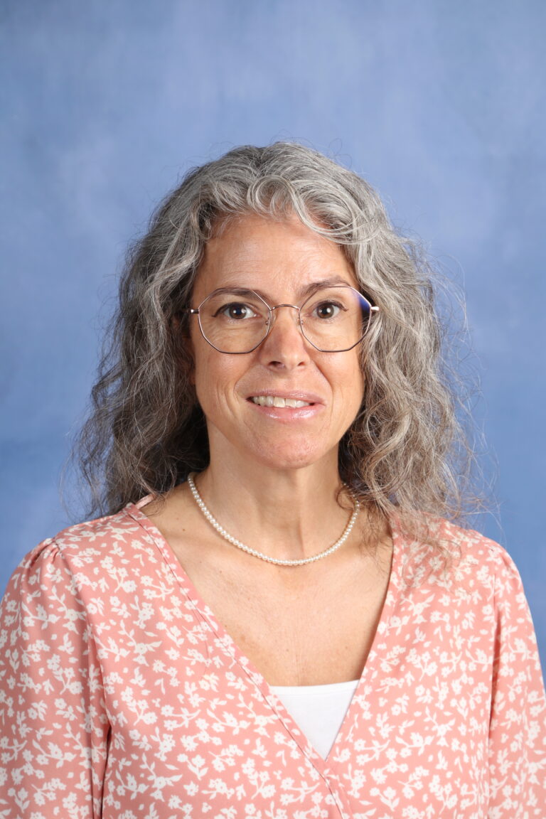 Portrait of Carlie Watt, wearing glasses and a floral top, against a blue background.