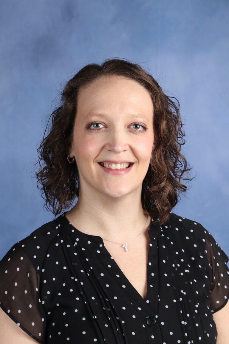 Emily Beck portrait. Woman with curly brown hair smiling in a black polka dot blouse.